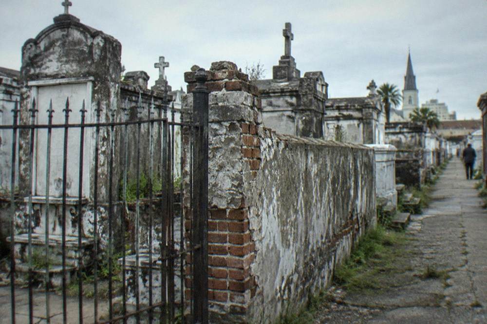 An iron gate and stone wall enclosing an old New Orleans cemetery with tall tombs behind it