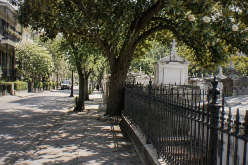 Sunlit Garden District street with the closed cemetery gates and tombs visible behind fencing