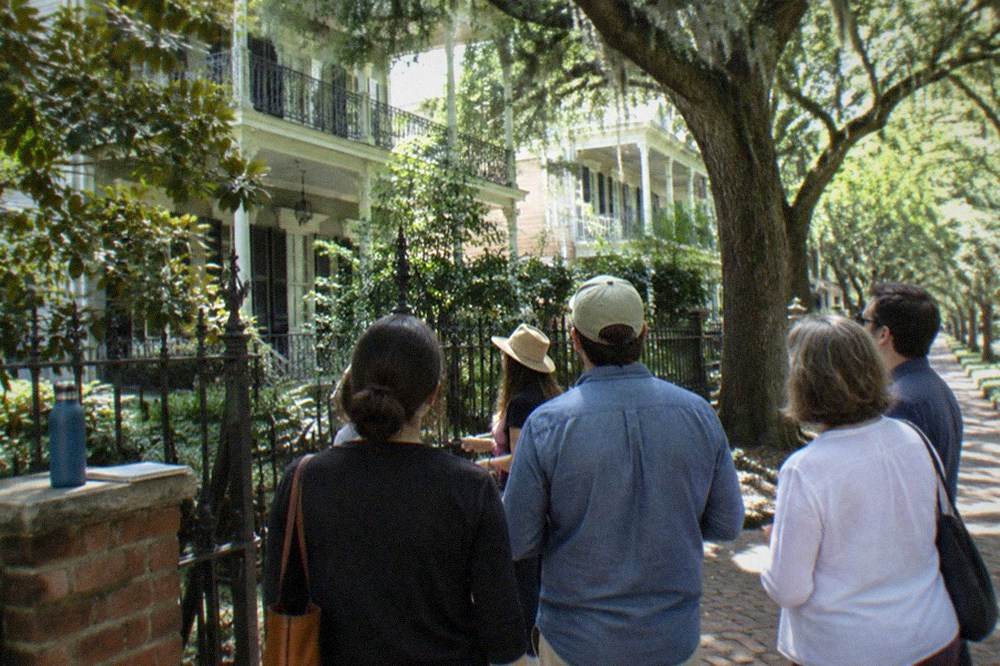 Preservation-style guided group gathered at a cemetery gate on a leafy New Orleans street