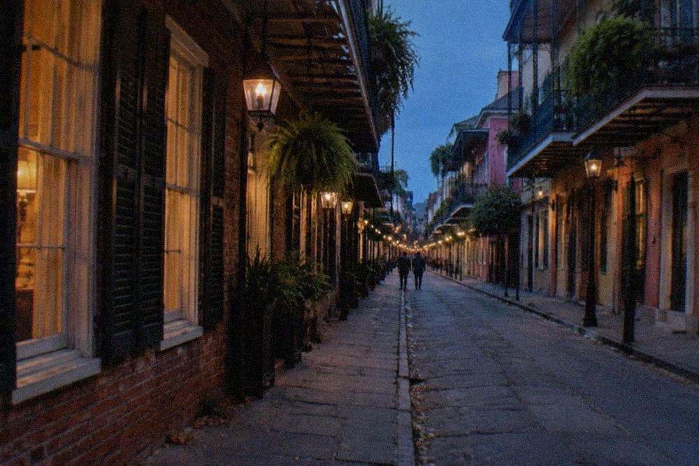 Narrow French Quarter street at dusk with lit windows and hanging lanterns above old brick buildings