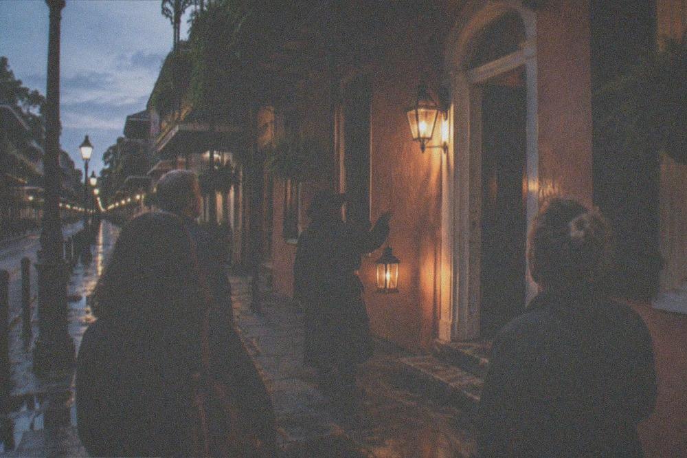Guide holding a lantern outside a lit French Quarter building as a small group gathers at dusk