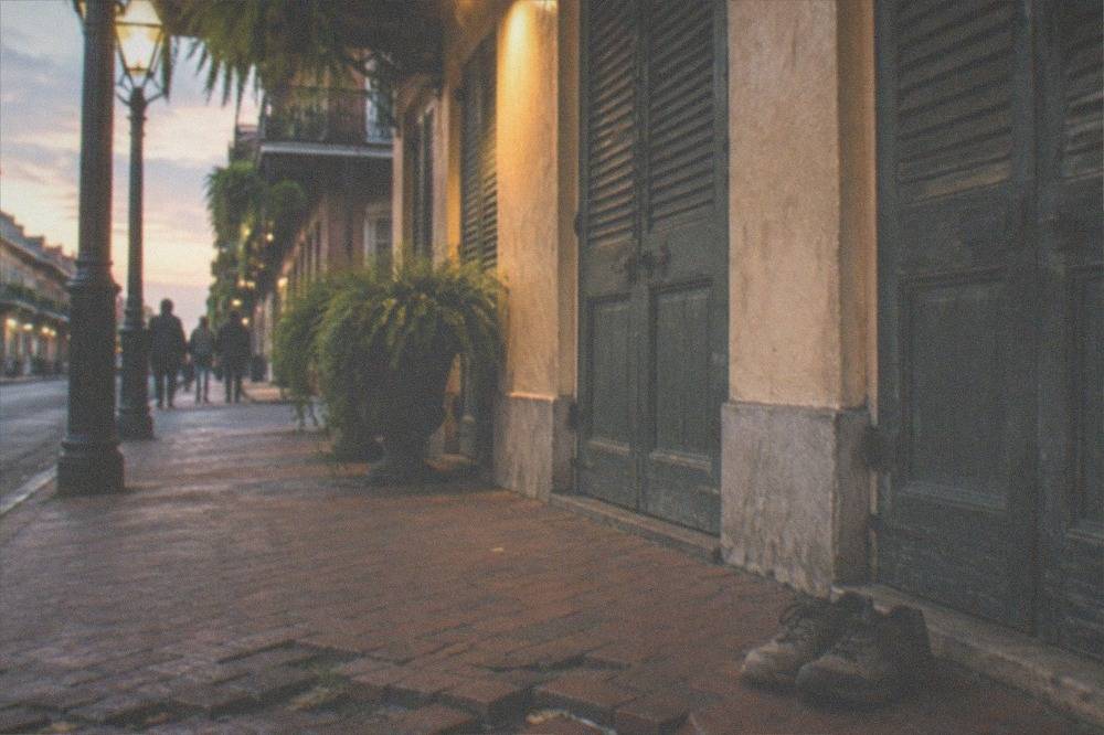 A pair of worn comfortable walking shoes resting on an uneven brick sidewalk beside a shuttered French Quarter storefront