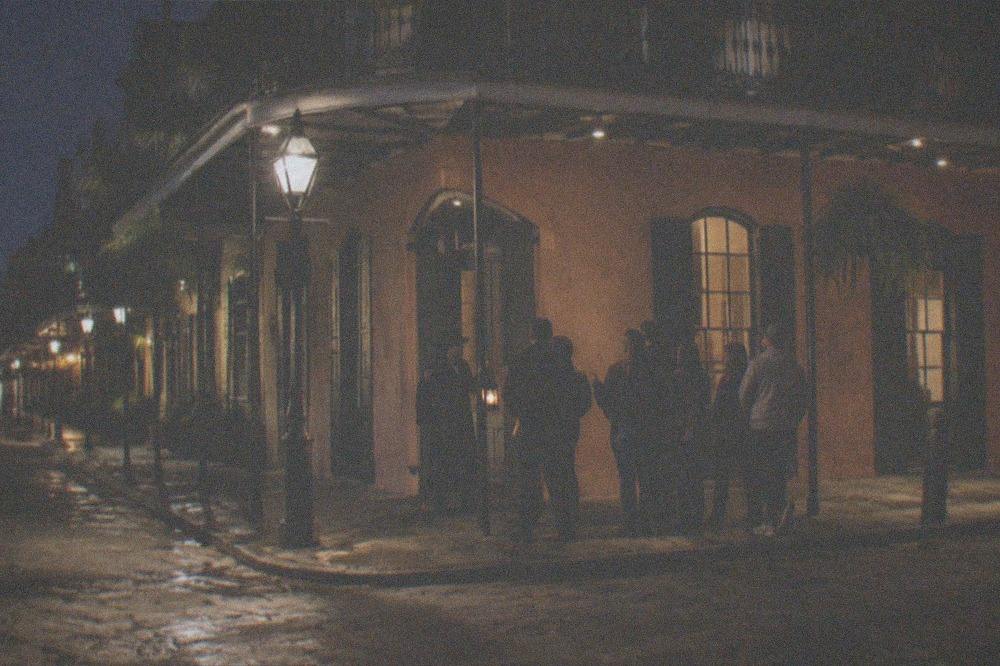 A licensed tour guide holding a small lantern addressing a small standing group on a lamplit French Quarter corner